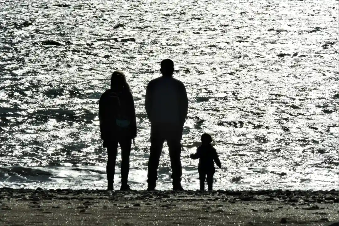Sylwetki dwojga dorosłych i małego dziecka stoją razem na kamienistej plaży, zwrócone ku jasnym, migoczącym falom – w scenerii pełnej spokoju i zadumy.