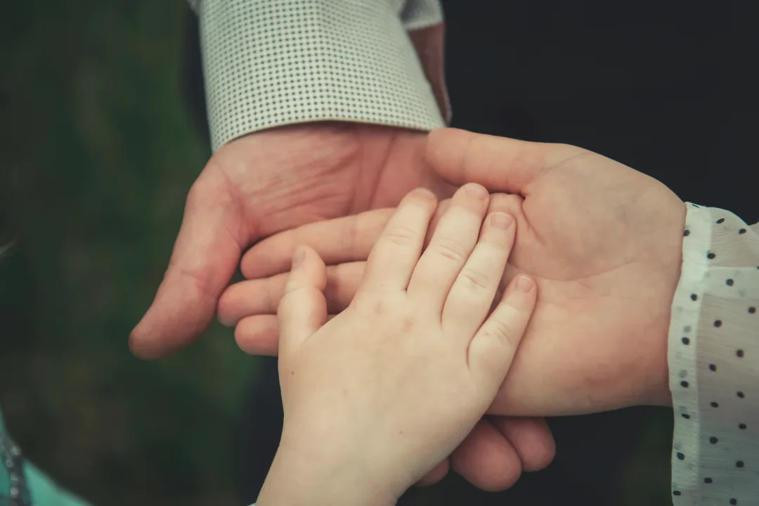 Mała dłoń dziecka spoczywa delikatnie na otwartej dłoni dorosłego – to czułe zbliżenie na rozmytym, zielonym tle, wyrażające troskę i poczucie bezpieczeństwa.